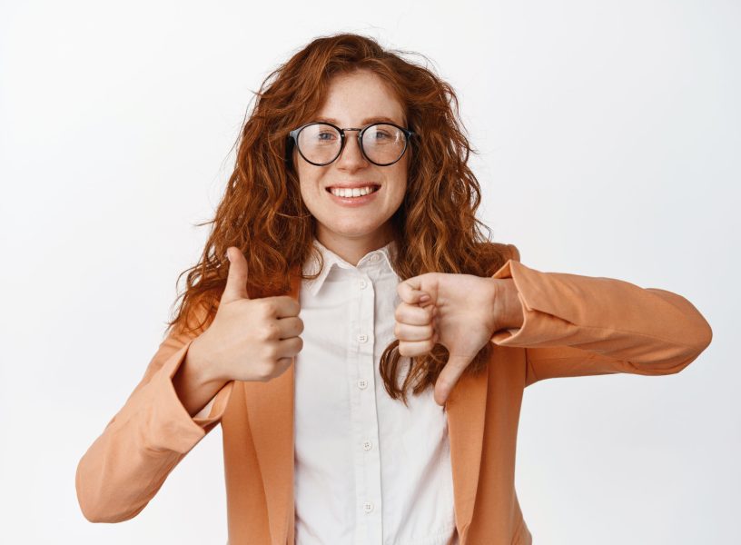 Like or dislike. Smiling saleswoman with red curly hair, showing thumbs up and thumbs down, advantage or disadvantage gesture, standing in suit and glasses over white background Like or dislike. Smiling saleswoman with red curly hair