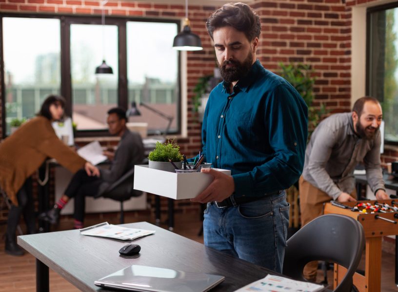Sad dismissed man holding box with belongings after being fired while his collegues looking at hom working in startup office. Businesspeople brainstorming ideas developing marketing project